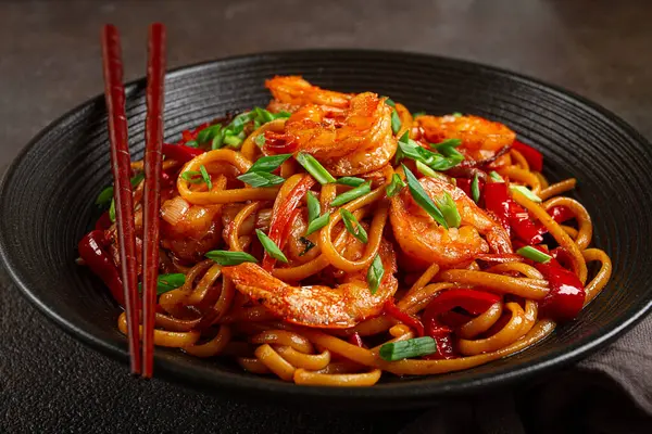 Kırmızı dolmalık biber ve yeşil soğanlı udon kızartmalı karides eriştesi, siyah bir kasede servis ediliyor, çubuklarla servis ediliyor, koyu arka plan...,