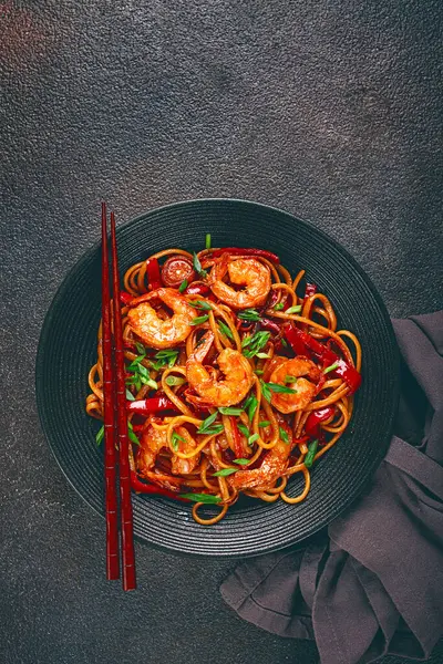 Kırmızı dolmalık biber ve yeşil soğanlı udon kızartmalı karides eriştesi, siyah bir kasede servis ediliyor, çubuklarla servis ediliyor, koyu arka plan...,