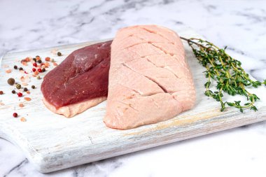 Taze çiğ ördek göğsü, ahşap bir kesim tahtasına yayılmış, beyaz mermer bir yüzey üzerinde otlar ve baharatlarla,