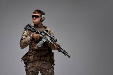 Tam teçhizatlı bir taktik operatörü, kamuflaj, eldiven ve koruyucu kulaklıklar takmış, profesyonelliği, hazır olmayı ve modern dövüş yeteneklerini tasvir ediyor.