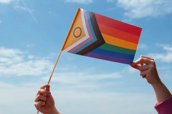 Two hands holding a flag with the new colors of the lgtb collective over a blue sky.