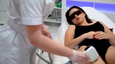 Koruyucu gözlüklü seksi kadın tıbbi kanepede uzanmış gülümserken kadın kuaför güzellik salonunda lazer tüy dökücü tüy döküyor. Güzellik epilasyonu kozmetoloji ve spa konsepti