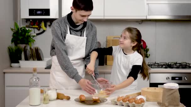 快乐而微笑的女孩用金属丝搅拌器在碗里搅拌鸡蛋 姐姐在厨房帮助哥哥 青少年做饭的过程饼干和糕点 紧密相连的家庭和幸福的童年 — 图库视频影像