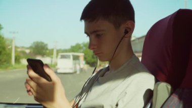Çocuk Avrupa seyahati sırasında toplu taşıma otobüsünde pencerenin yanında otururken akıllı telefon kullanıyor. Online oyun oynuyorum. Mesaj yazar. Telli kulaklıkla müzik dinliyor.