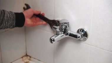 Erkek Tesisatçı su sızdıran banyoyu tamir ediyor. Su musluğunu ingiliz anahtarıyla tamir ediyor. Evde tanınmayan bir adam çalışıyor. Duvar kiremitlerinde küf olan eski banyoda tesisat tamiri.