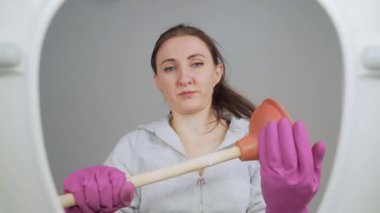 Odaklanmış kadın, pompayı kendine güvenerek elinde tutarken su tesisatı sorunlarıyla uğraşmaya hazır. Tuvalet temizliği için hazırlanmış mor lastik eldivenli bir kadın. Tuvaletin dibinden bak. Temizlik