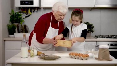 Şefkatli büyükanne, torununa sıcak beyaz mutfakta hamur için un elemeyi öğretiyor. Pasta için süt yumurtalı, ayçiçeği yağlı ev yapımı kekler. Yaşlı bir kadın, küçük bir kızla birlikte süzgeçten un döker. Pişirme