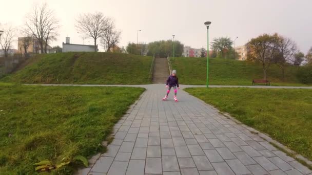 膝と肘の安全パッドの小さな女の子は 市立公園の屋外でローラーのスケートに乗ることを学びます アクティブなスポーツ 幅広い視野を楽しむキッズ 市立公園に沿ってローラーブレードする陽気な注意なしの若い少女 — ストック動画