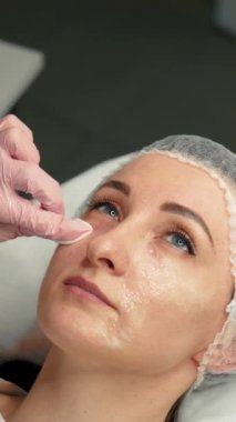 Kozmetik uzmanı, kadınların ciltlerini yumuşatmak, ciltlerini yumuşatmak ve nemlendirmek gibi estetik ameliyatlardan sonra kadınların yüzünü pamuklu süngerle siliyor. Dikey oryantasyon. Gençleştirme