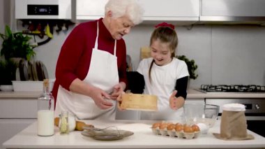 Küçük kız gülümseyip unu elek temizliyor. Büyükanne ve torun mutfakta pizza hamuru hazırlıyorlar. Evde pişireceğim. Mutlu çocuk büyükannesiyle iyi vakit geçirdiği için gülümsüyor.
