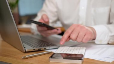 Adam dizüstü bilgisayarla masada oturuyor. Hesap makinesi ve cep telefonu kullanarak masrafları hesaplıyor. Görüntüyü kapat. İş adamı harcamaları yönetir, kişisel harcamaları kontrol eder. Uzak iş için dizüstü bilgisayar kullanan erkek yönetici