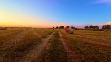 Sunset 'teki saman balyaları. POV traktörü kırsal kesimden geçiyor. Saman balyalarıyla sahanın üzerinde güzel bir gün batımı. Toprak yol tarımın ortasından geçiyor.