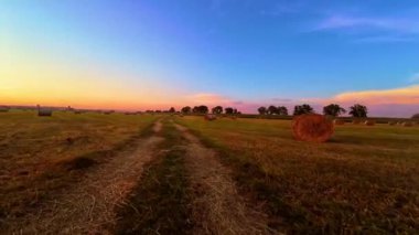 Kırsal bölgedeki Sunset 'te Hay Bales ve Dirt Road. POV traktörü kırsal kesimden geçiyor. Kirli yol kırsal alanda gün batımında düzgünce istiflenmiş saman balyalarıyla kaplı. Altın saat