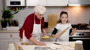 Neşeli kız ve büyükannem hamurla turta pişiriyorlar. Çocuk dans ediyor ve mutfakta eğleniyor. Evde kahvaltı için yemek pişirmek. Mutlu bir çocuk ve mutfakta yaşlı bir kadın.