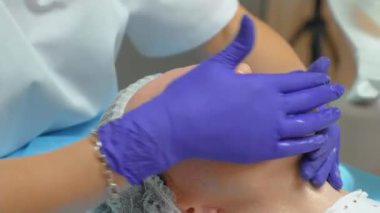 Güzellik merkezindeki ameliyatlardan önce kozmetolojist hastanın yüzünü yıkıyor. Yüz derisi, yakın çekim. Doktor, kozmetik ürünlerini mutlu bir kadının yüzündeki temizleme jeliyle tamamen yıkar.