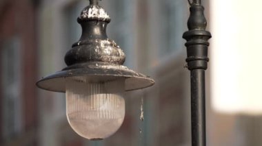 Arka plandaki binalarla Sunny Urban Environment 'ta Lamppost' ta asılı Rustic Sokak Lambası. Vintage sokak lambası, güneş ışığıyla aydınlatılan sokak lambasının üzerine konur.