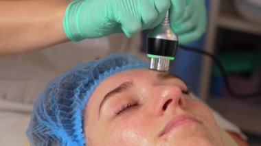 Yüzün donanım kozmetolojisi yakın plan. Profesyonel nemlendirici ve gençleştirici. Eldivenli güzellik uzmanı, kozmetoloji kliniğindeki genç bayanın ultrasonik oyuk yüz hatlarını düzenliyor.