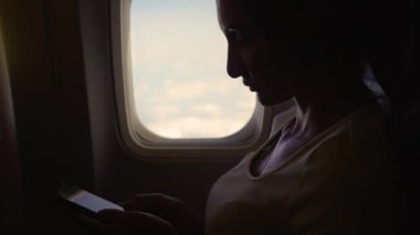 Genç kadın gündüz uçuşu sırasında pencerede otururken akıllı telefonunu kullanıyor. Uçak penceresinin yanında oturan genç ve güzel bir bayan, akıllı telefonunu uçakta tutuyor.