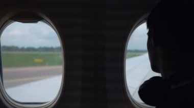 Havalimanında kalkışa hazırlanırken uçak camından dışarı bakan kulaklık takan bir genç. Genç çocuk uçak penceresinden dışarı bakıyor, kalkışı bekliyor.