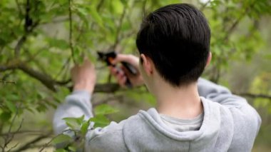 Bahçede çalışan genç bir çocuk budama makası ile ağaç dallarını buduyor. Ergen çocuk bahçedeki ağaçların dallarını kesmek için budama aleti kullanıyor. Küçük çocuk ağaç dallarını bahçe aletleriyle buduyor.