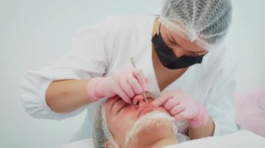 Beyaz elbiseli ve pembe eldivenli kadın sivilcelerini müşteri yüzünden çıkarıyor. Tedavi sırasında metal alet kullanan kadın hasta tenine odaklanıyor. Tıbbi maske takan ve saç filesi takan bir kız.