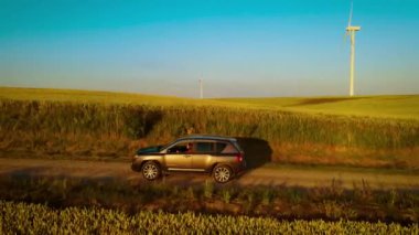 Yeşil buğday tarlaları ve uzaktan görülebilen rüzgar türbinleriyle çevrili, toprak yolda ilerleyen bir araba. Kadın sürücü sakin bir akşam manzarasının tadını çıkarırken kırsal arazide süzülen SUV