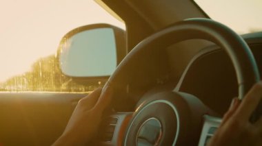 Gün batımında direksiyonda direksiyon tutan tanınmayan eller. Parmaklar, altın gece ışığında direksiyonda gezinen araçlar. Sürücü sıcak günbatımıyla çevrili huzurlu yolculuğun tadını çıkarıyor