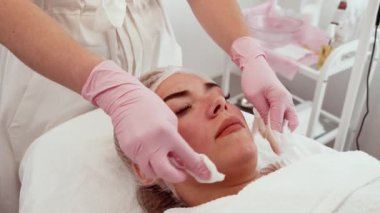 Kadın tedavi masasında rahat bir ifadeyle yatıyor, profesyonel temizlikçi yüzü ve pamuk pedleri. Cilt bakımı sırasında kadın istirahati, uzman tarafından yapılan nazik temizlik. Bayan dinleniyor