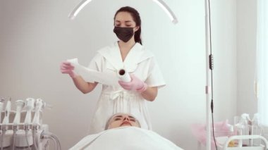 Kozmetik bilimci kozmetoloji kliniğinde prosedür hazırlıkları sırasında tek kullanımlık kağıt rulosu tutuyor. Yüz bakımı için eldiven ve maske takan kadın uzman.