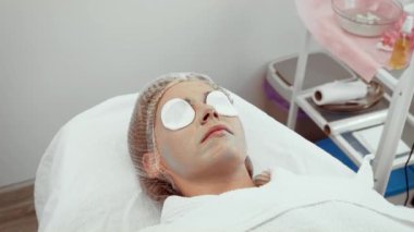 Müşteri estetik merkezinde canlandırıcı yüz tedavisi görüyor. Besleyici maskeli bir kadın dinleniyor, cilt tazelemeyi teşvik ediyor. Pamuk pedler seans sırasında rahatlatıcı etkileri artırır