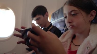 Genç kız ve erkek uçuş sırasında uçakta seyahat ederken akıllı telefondan oyun oynamanın keyfini çıkarıyorlar. Genç bir çocuk ve kız, ebeveynleri olmadan uçakla seyahat ederken cep telefonuyla oyun oynuyorlar.