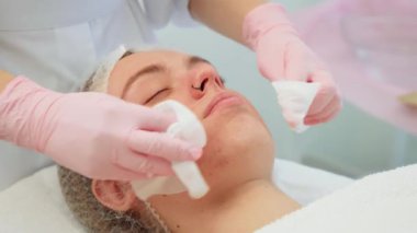 Eldivenli ellerle pamuk pedlerle yüzünü temizlerken tedavi masasında uzanan sakin yüzlü bir kadın. Kadın güzellik prosedüründen önce steril ortamda yüz hazırlığı yapıyor. Hanımefendi.