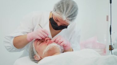 Beyaz elbiseli ve pembe eldivenli bir kadın müşteriye yüz güzelliği ameliyatı yapıyor. Kadın tedavide metal aletleri dikkatlice kullanırken tenine odaklanıyor. Tıbbi maske takan ve saç filesi takan kız.