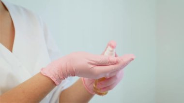 Cilt bakımı sırasında elinde sıvı sprey antiseptik tutan kadın. Pembe eldiven giyen ve dezenfektan kullanmaya hazırlanan bir kadın. Klinik aydınlatma altında şişe ile ellerinizi yakın çekim
