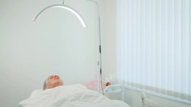 Kozmetik kliniğinde, kavisli ışık altında tedavi masasında yatan bir kadın. Profesyonel ekipmanlarla cilt bakımı sırasında kadın dinleniyor. Parlak klinik ortamında güzellik tedavisi gören kız