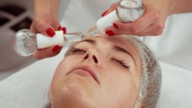 Cilt bakımı kliniğinde kadın alnında cam küreler kullanan kozmetolog, rahatlama ve cilt tazeleme üzerine odaklanıyor. Kadın sakince yatıyor, kan dolaşımını hızlandırmak için soğutma terapisinin tadını çıkarıyor.