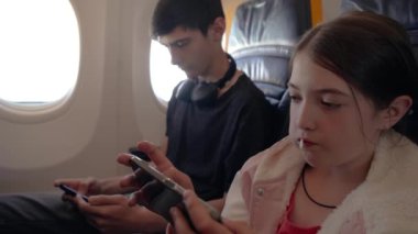 Genç Adam ve Kız Yalnız Seyahat Ederken Cep Telefonlarında Oyun Oynamak İçin Kulaklık Kullanıyor. Çocuklar uçakta oturur, akıllı telefonlarına odaklanır, uçarken oyun deneyiminin tadını çıkarırlar.