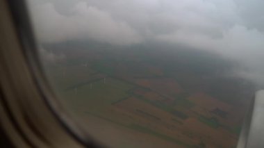 Alçak Bulutlar Arasında Rüzgar Türbinleri İniş İçin Hazırlanan Uçak. Pencerenin yanında türbin ve kanatlar var. Kabarık beyaz bulutların üzerinden geçen uçak