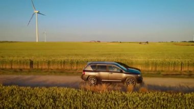 Sıcak akşam güneşi altında yeşil buğday tarlaları olan toprak yolda SUV kullanan bir kadın. Dişi, kırsal alanda sakin manzaranın tadını çıkarırken kırsal alanda araba sürüyor. Otomatik olarak gezen kadın manzaralı yolda