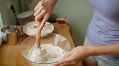 Kadın, un ve hamuru pişirme işlemi sırasında tahta spatula kullanarak cam kasede karıştırıyor. Dişi, pürüzsüz hamur yaratmak için kuru ve ıslak malzemeleri dikkatlice katlıyor. Bayan yemek tarifi hazırlıyor.