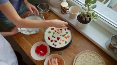 Anne, kız ve oğul kremalı pasta dolgusuna jöle parçaları koyuyorlar. Kadın, kız ve erkek krem şanti tabakasında renkli jelatin küpleri düzenliyorlar. Bayan, çocuk ve çocuğun tatlıyı süslemesi.