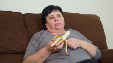 Yetişkin bir kadın kanepede oturmuş muz yiyor. Son sınıf dişisi soyulmuş meyveyi tutarken dinleniyor. Bayan kanepede dinlenirken atıştırmanın tadını çıkarıyor.