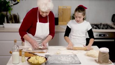 Büyükanne ve torun. Evde eğlence, öğrenme ve çocukluk gelişimi için mutfakta pişirmek. Un ve hamurla yuvarlanan raptiyelerle sevimli kıza yemek pişirmeyi öğreten mutlu büyükanne.