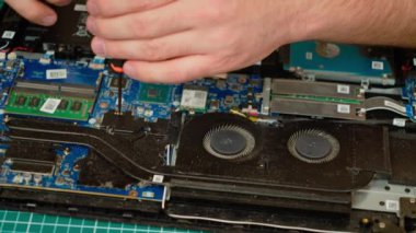 Tanınmayan bir adam dizüstü bilgisayarını tornavidayla onarıyor. Erkek, tozlu cihazın iç bileşenleri üzerinde çalışıyor. Eleman elektronik devre ve soğutma sisteminde bakım yapıyor.