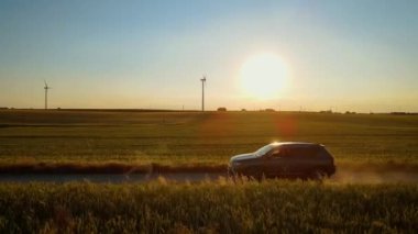 Çamurlu yolda araba sürüyor. Kadın direksiyonda. Arka planda rüzgar türbinleri ve toz bulutları. Kadın sürücü, güneş ışığı altında kırsal manzaranın tadını çıkarırken SUV yolda ilerliyor.