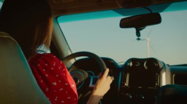 Kendine güveni tam bir kadın, direksiyonu dikkatle ve titizlikle tutuyor. Şık bir SUV, arka planda altın güneş ışığı ve rüzgar türbinleri ile çerçevelenmiş şekilde kırsalda hareket eder. Kararlı kadın.