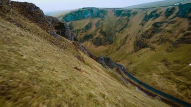 Etrafı sarp çim kayalıklarıyla çevrili virajlı dağ yolu boyunca araba sürüyor. Araç İngiltere 'nin Peak District bölgesinden manzaralı bir yerden geçiyor. Kırmızı araba virajlı asfalt yolda ilerliyor.