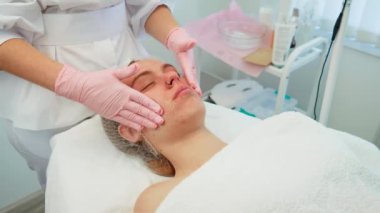 Eldivenli ellerle yüzüne özenle masaj yaparken sakince uzanan kadın. Kadın kozmetoloji seansı sırasında yüz tedavisi görüyor. Uzman olarak dinlenen bayan cilt bakımını nazikçe yapar.