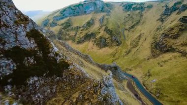 Sarmal dağ yolundan geçen araba dik çim kayalıklarıyla çevrili. Araç, Birleşik Krallık 'ın Peak District bölgesinde manzaralı bir yolda ilerliyor. Kırmızı araba virajlı asfalt yolda engebeli bir yolda ilerliyor.