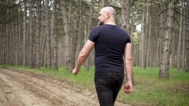 Dar tişörtle çam ormanında yürüyen kel sakallı bir adam. İnce gömlekli, yeşil ağaçların arasında gezinen bir erkek. Etrafı çam ağaçlarıyla çevrili ağaçlık yolda yürüyen bir adam.